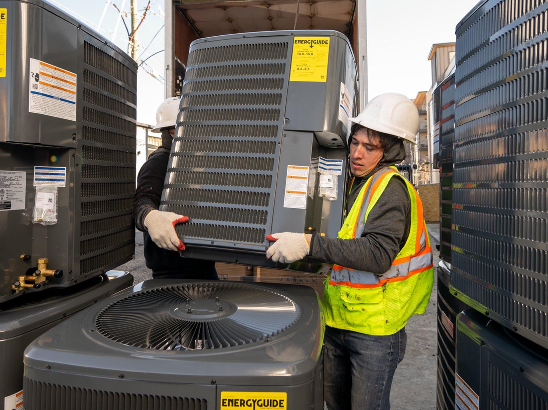 Two workers wearing PPE carry an air conditioner condenser.